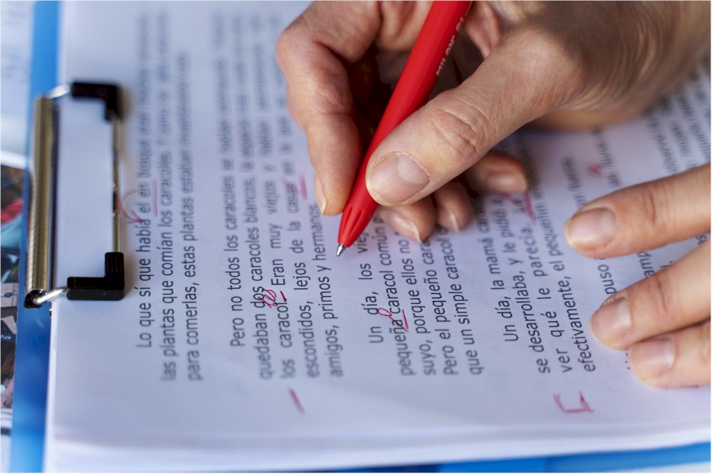 Photo of a hand holding a pen. It's owner is correcting text on a clipboard in red ink - Image by freepik